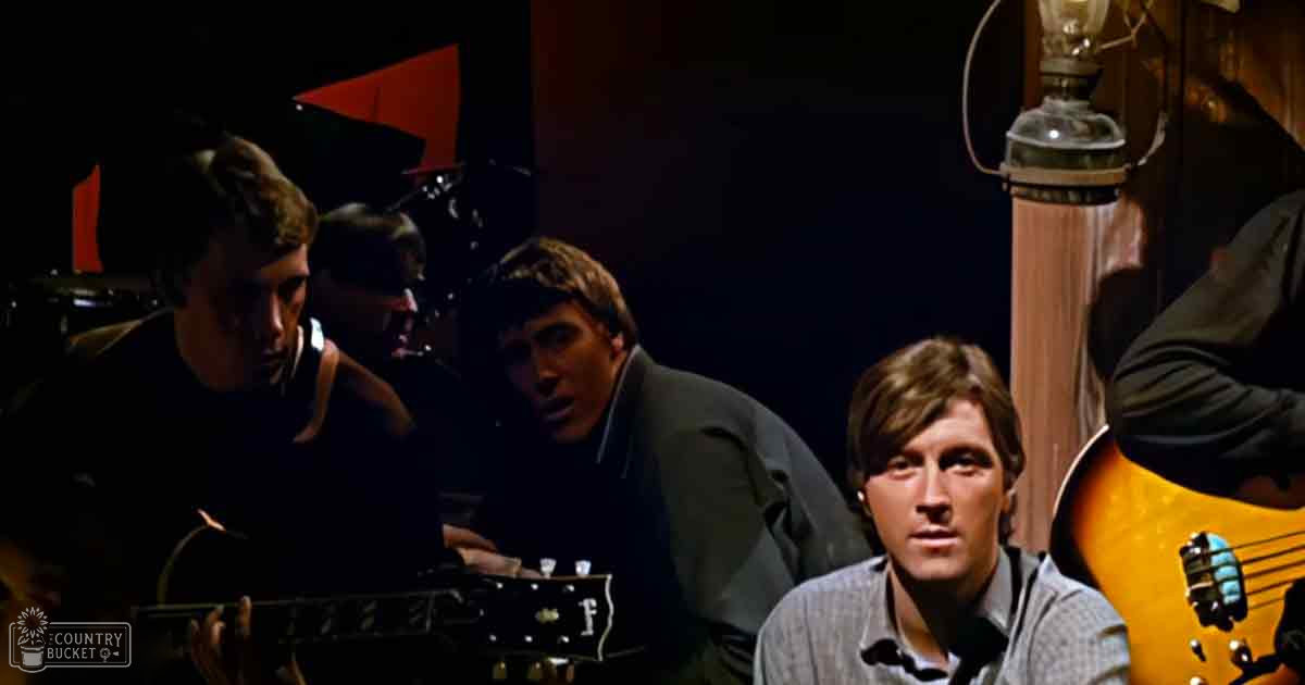 Indoors in a music studio with young adult males wearing formal or casual attire, holding musical instruments under artificial evening lighting.