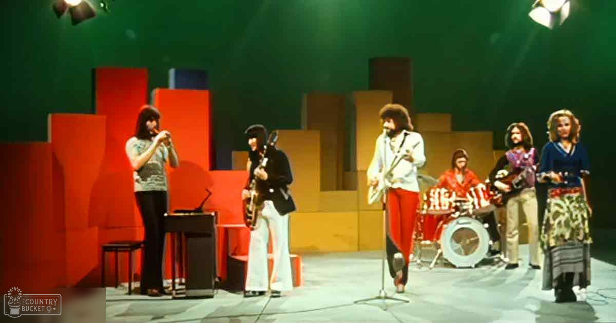 An indoor music studio with green screen backdrop, with young adult females in patterned tops and pants alongside adults in varied outfits, including a male drummer wearing red; setting suggests controlled lighting typical of recording environments.