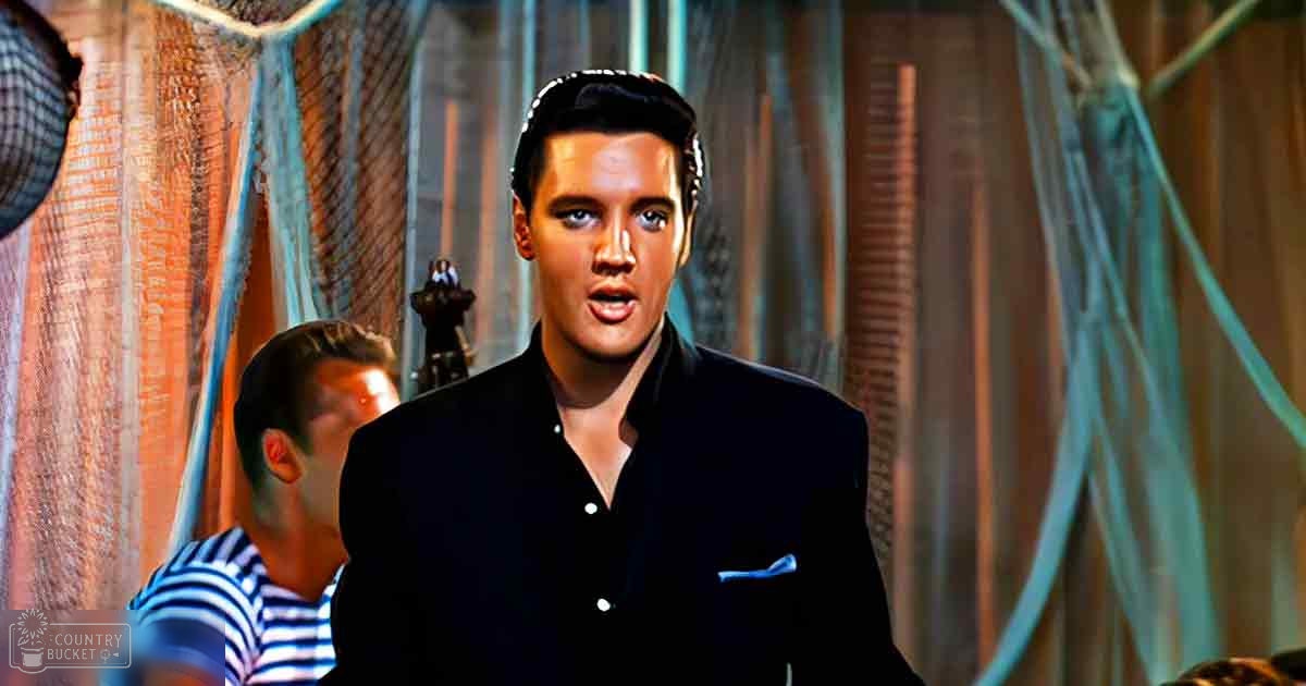 Young adult male wearing dark suit jacket and pocket square stands on stage with microphone visible behind him; background suggests theatrical setting with textured curtain backdrop.