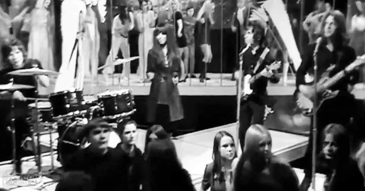 A musical performance indoors with a child in the foreground wearing a dark jacket, an adult female on stage in a dark coat and bob haircut, and mannequins dressed in formal attire in the background.