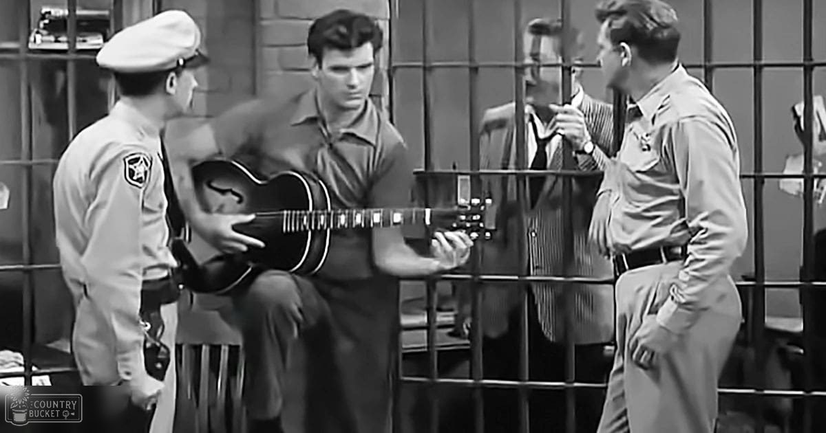 A man playing guitar inside a jail cell with uniformed officers nearby.
