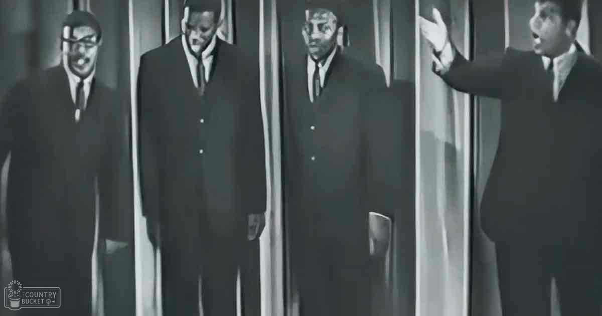 Group of men dressed in dark suits, possibly from a performance or music video setting; indoor backdrop suggests formal recording environment.