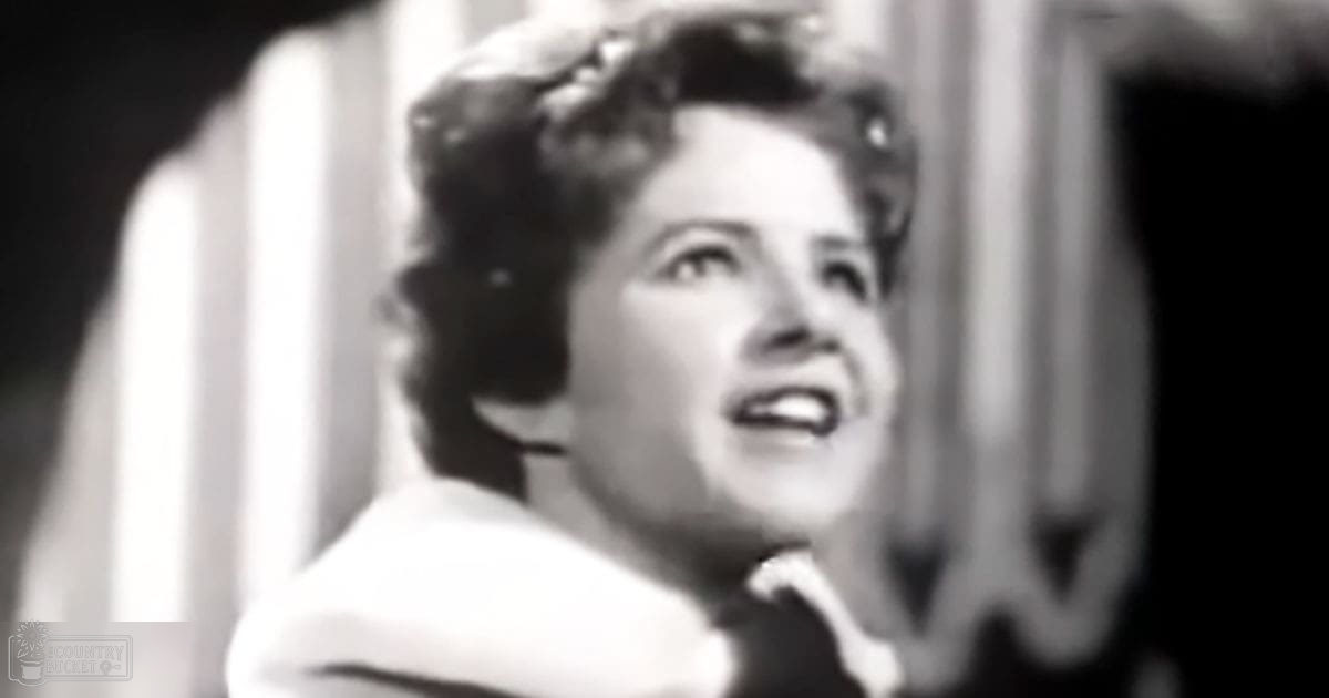 An adult female in a formal, light-colored jacket with high collar indoors, ornate background suggests performance or recording setting