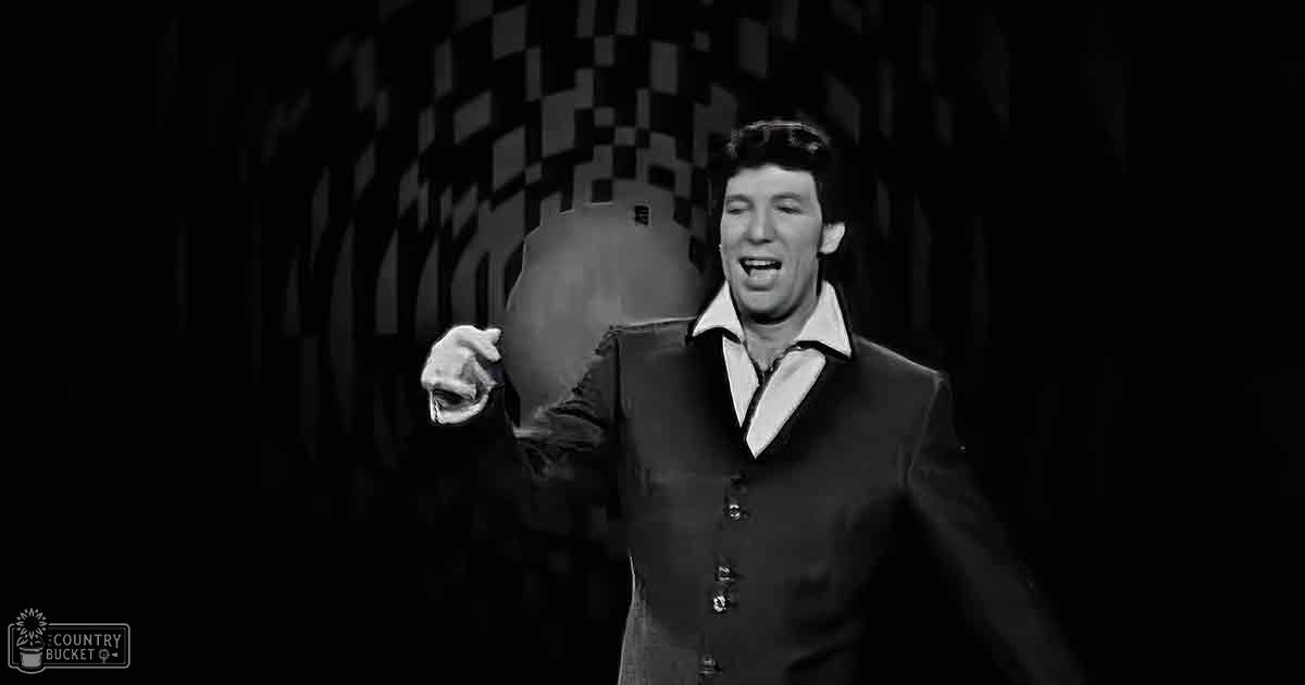 Adult male performer in formal black suit jacket with buttons down the front, holding a microphone on stage under dimly lit stage lighting.