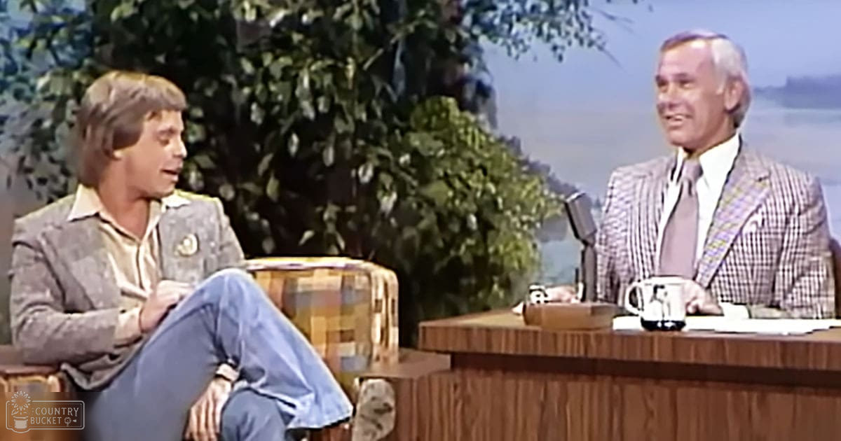 A young adult male in a patterned blazer and jeans sits beside an older adult male in a checkered suit jacket at a talk show set with greenery visible through windows, suggesting spring or summer.