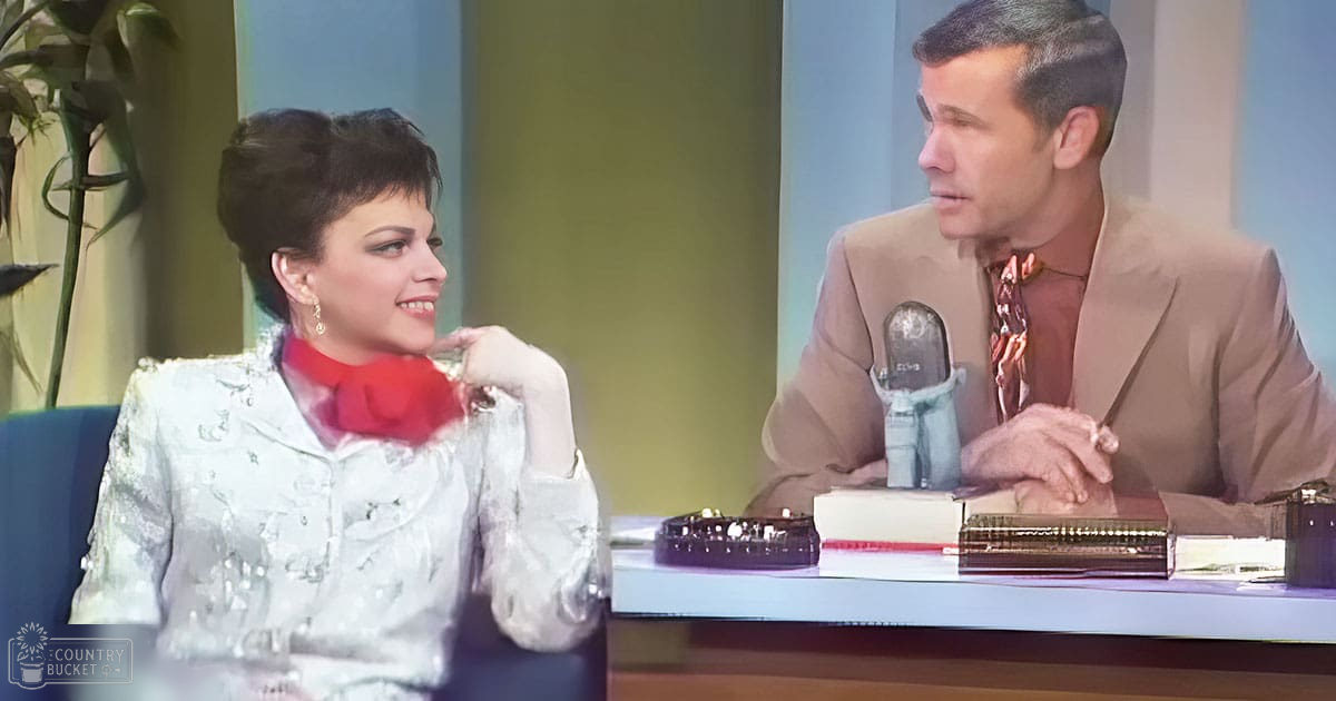 An adult female in a white sequined 1960s-style jacket with long sleeves and a high neckline, being interviewed by an adult male in a beige suit jacket and patterned tie. The setting is a studio with green walls and plants, suggesting an indoor talk show or interview environment.