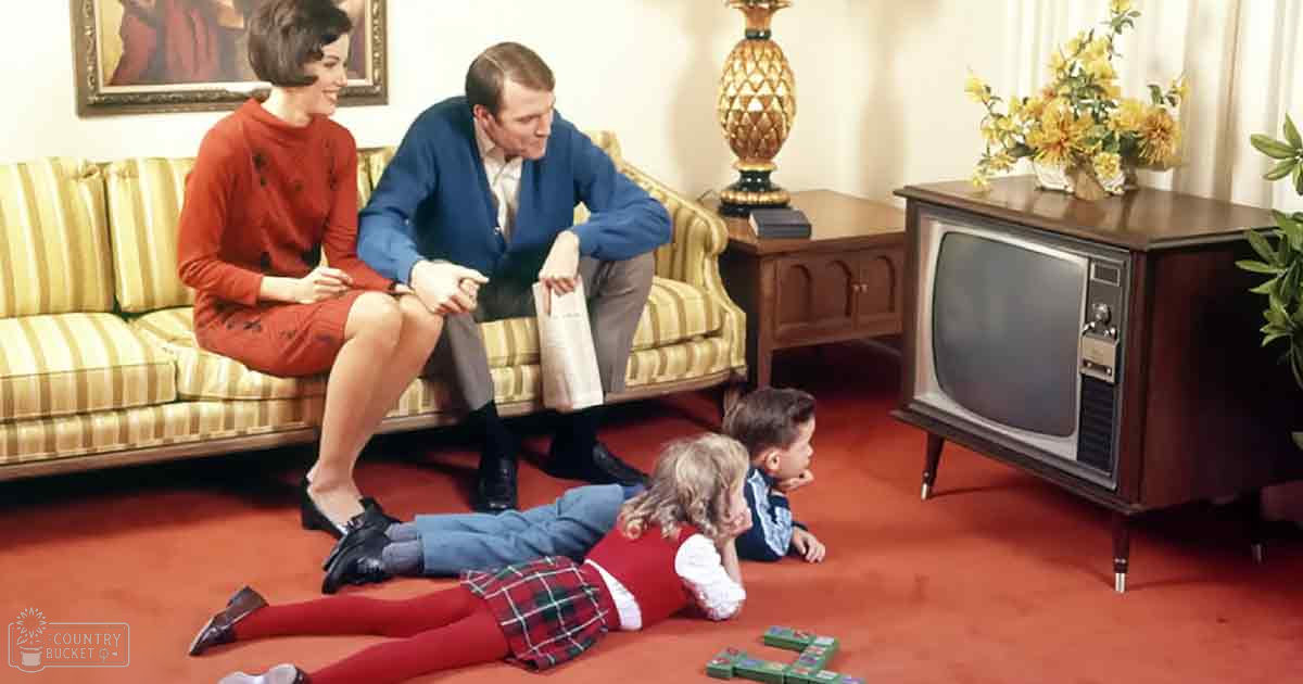 A family of adults and children gathered indoors on a striped yellow couch, wearing various patterns in reds, blues, and yellows under natural daytime light.