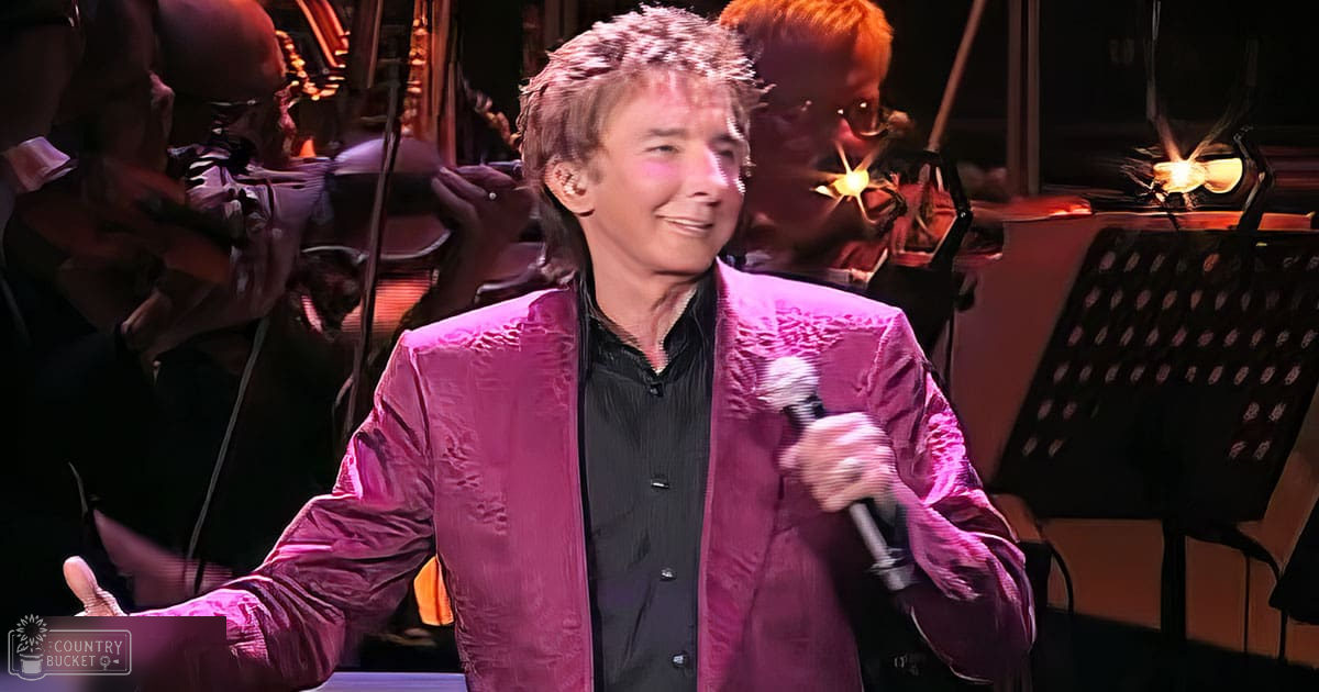 A middle-aged male performer in a shiny pink jacket with intricate embroidery, standing on stage during an indoor concert.