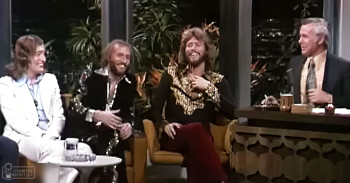 Adult males in a casual indoor setting, possibly a talk show, with greenery outside large windows and plants in the background.
