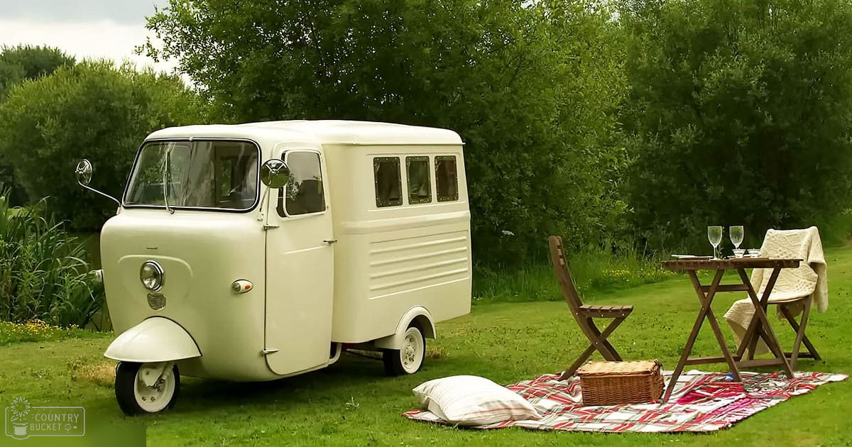 A vintage three-wheeled vehicle on a red-and-white checkered blanket in an outdoor park setting with lush green grass and trees, suggesting spring or summer.