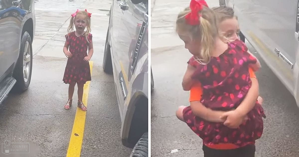 A toddler girl with a red polka dot dress and pink bow in her hair stands between two cars in an outdoor parking lot with wet pavement.