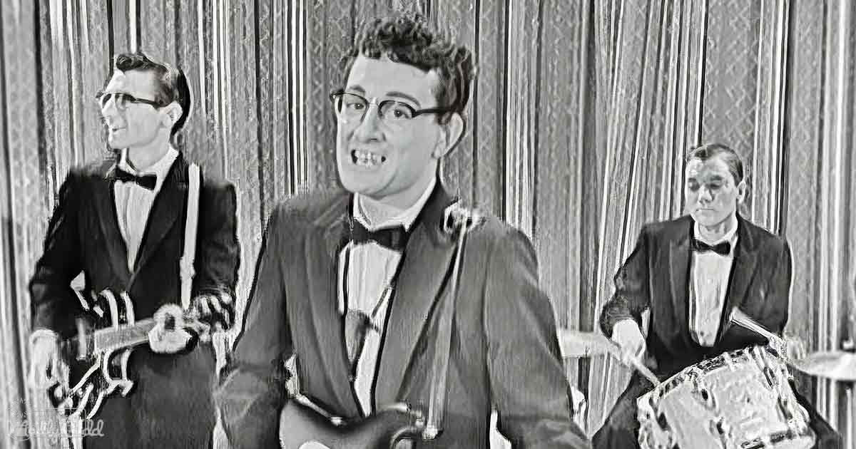 A young adult male singer wearing a dark suit, white shirt, and black bow tie stands in the center of an indoor performance setting with two other adults flanking him; one holding an electric guitar, another a drum set.