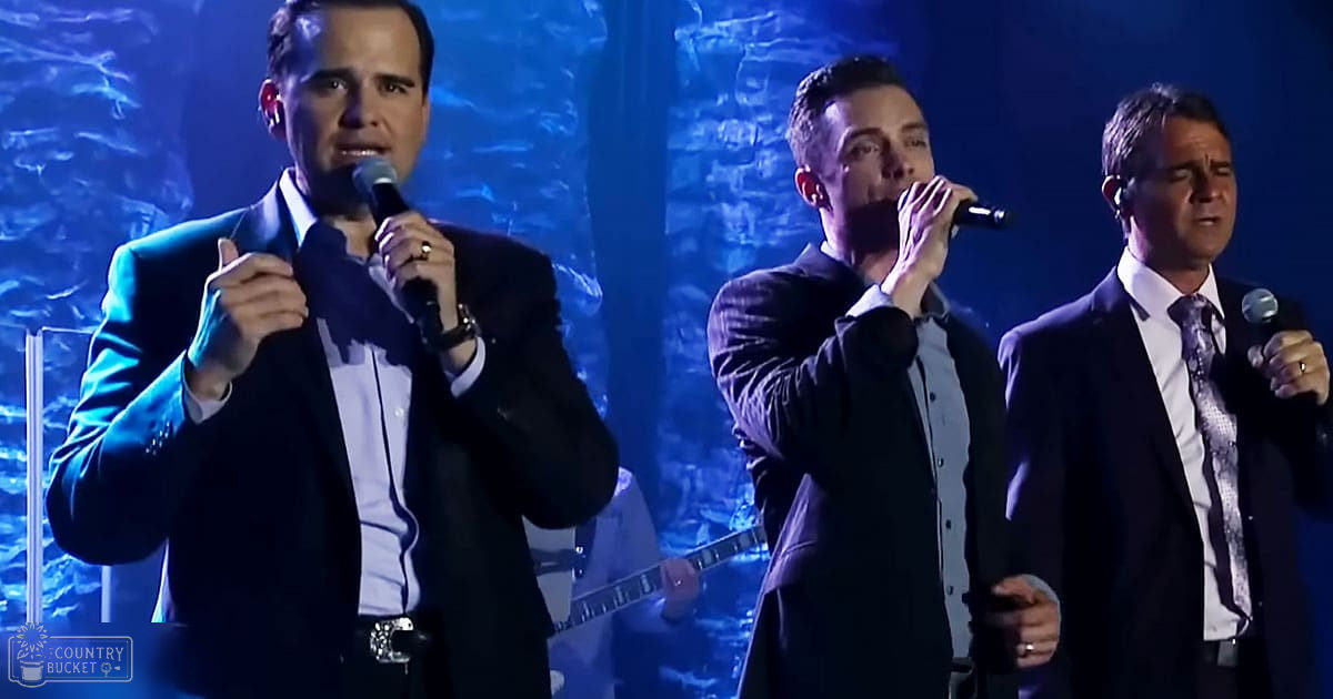 Three adult males singing on an indoor stage with blue lighting in a performance setting.
