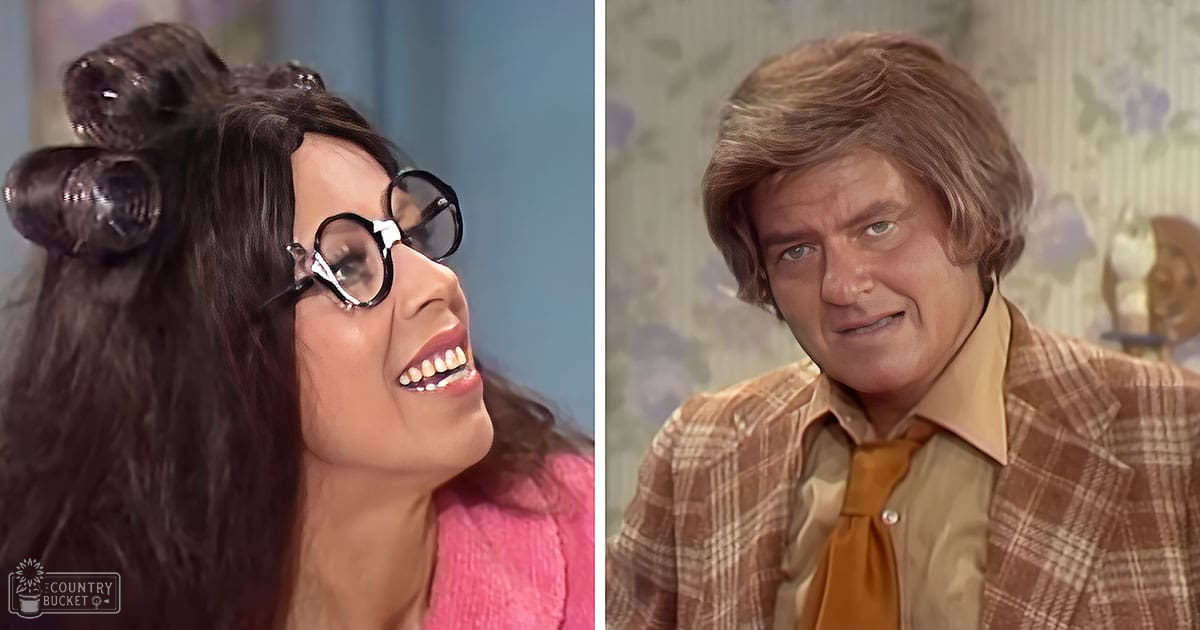 Young adult female wearing a pink sweater with large black-rimmed glasses, interacting with an older male middle-aged individual in a plaid jacket and yellow shirt in a floral-patterned living room setting during daytime.