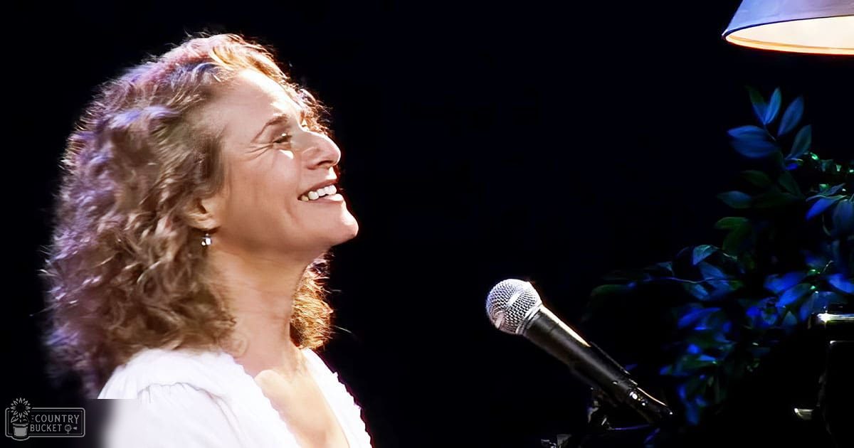 A middle-aged adult female in a white ruffled blouse with long sleeves and round neckline sings into a microphone against an indoor backdrop with a dark background, greenery, and a blue lampshade.