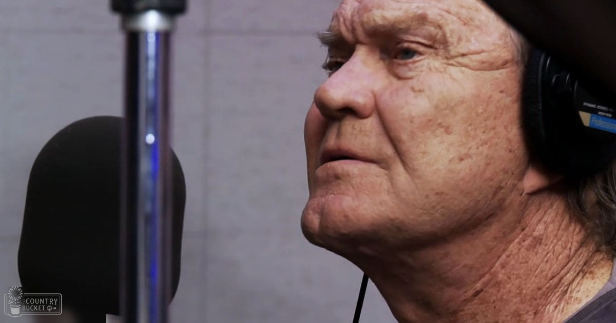 An older adult male in a recording studio with a dark hoodie, over-ear headphones, and acoustic foam panels visible behind him.