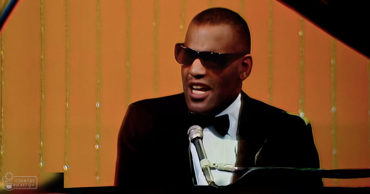 Adult male in a black suit jacket with a white shirt, wearing dark sunglasses, standing indoors against an orange wall with vertical gold stripes. The setting suggests a performance or presentation area.