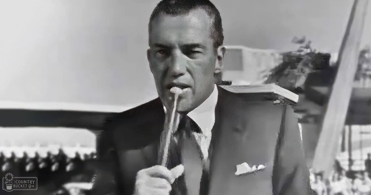 Adult male in a dark suit jacket with white pocket square near water body at an outdoor event during daylight hours.