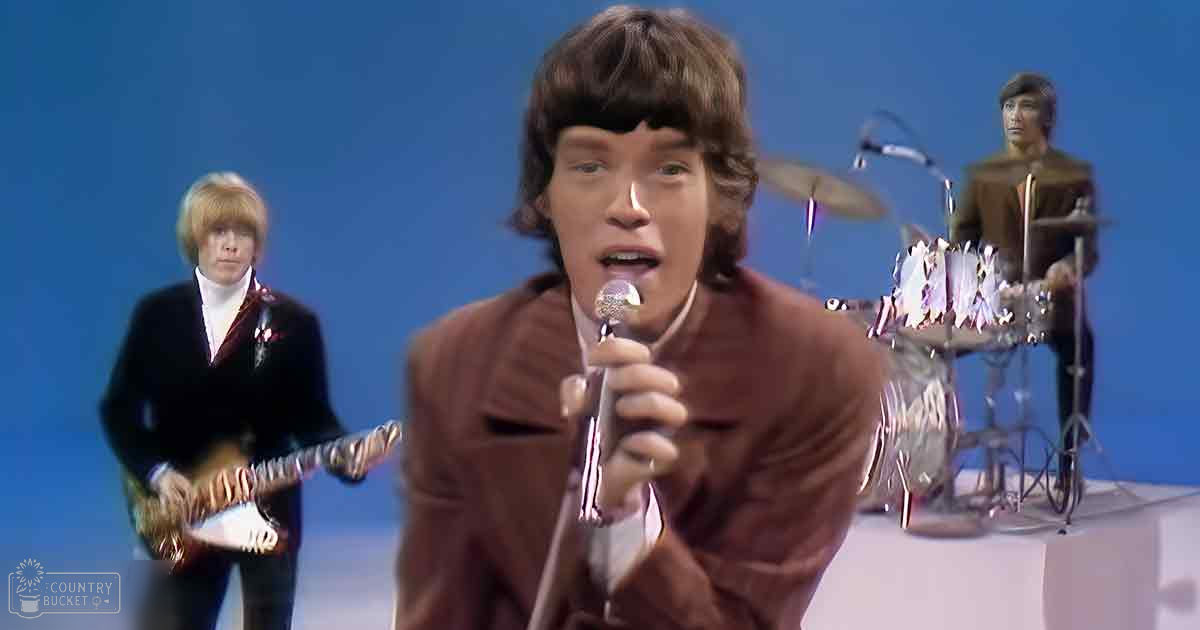 A young adult male singer in a brown suit jacket with two other musicians, one playing guitar and another drumming. The performance takes place against a solid blue background under artificial lighting.