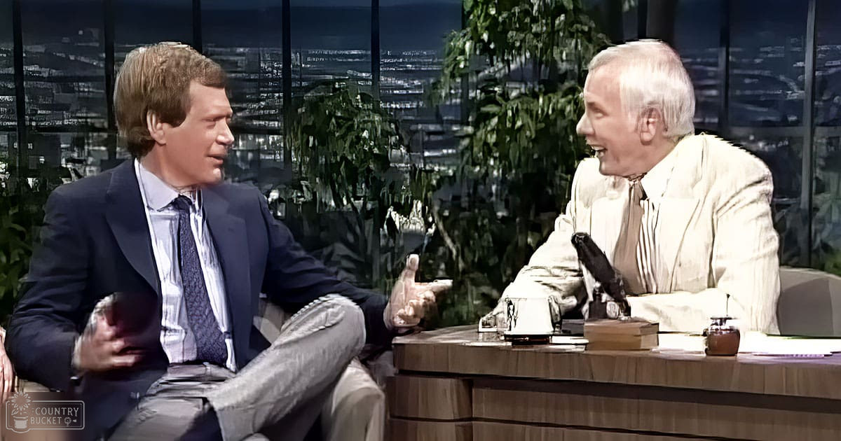 A talk show setting with two older adult males, one wearing a dark blue suit jacket and the other in a light-colored blazer, against a backdrop of an illuminated cityscape at night with greenery.