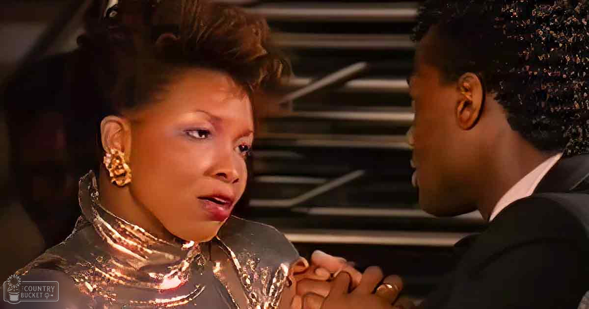 Adult female in a shiny metallic top and large hoop earrings, accompanied by an adult male in a dark suit jacket indoors with warm artificial lighting.
