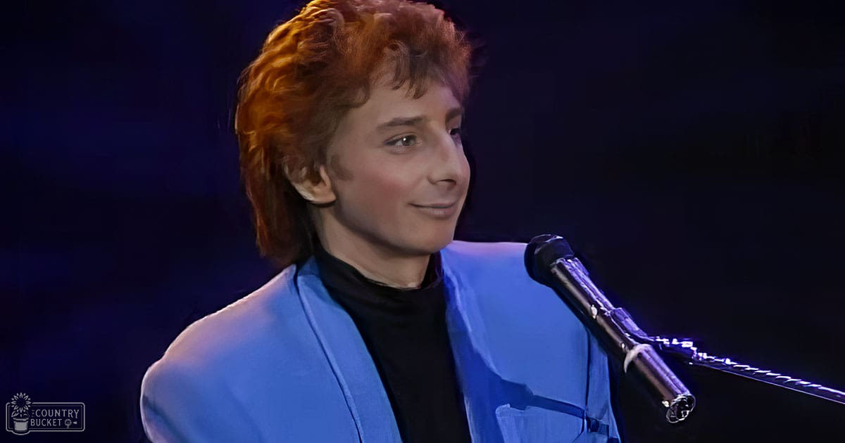 A young adult male stands indoors, wearing a blue blazer over a black turtleneck sweater. The setting suggests an event or performance venue with stage lighting.