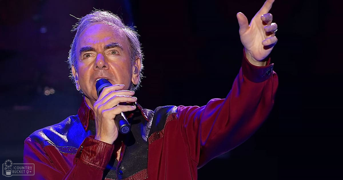 A middle-aged adult male singer in a fitted, shiny red jacket performing on an indoor concert stage with dramatic stage lighting.