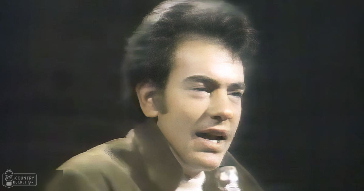 A young adult male wearing a dark-colored, collared, relaxed-fit shirt with short sleeves stands indoors under dim lighting.