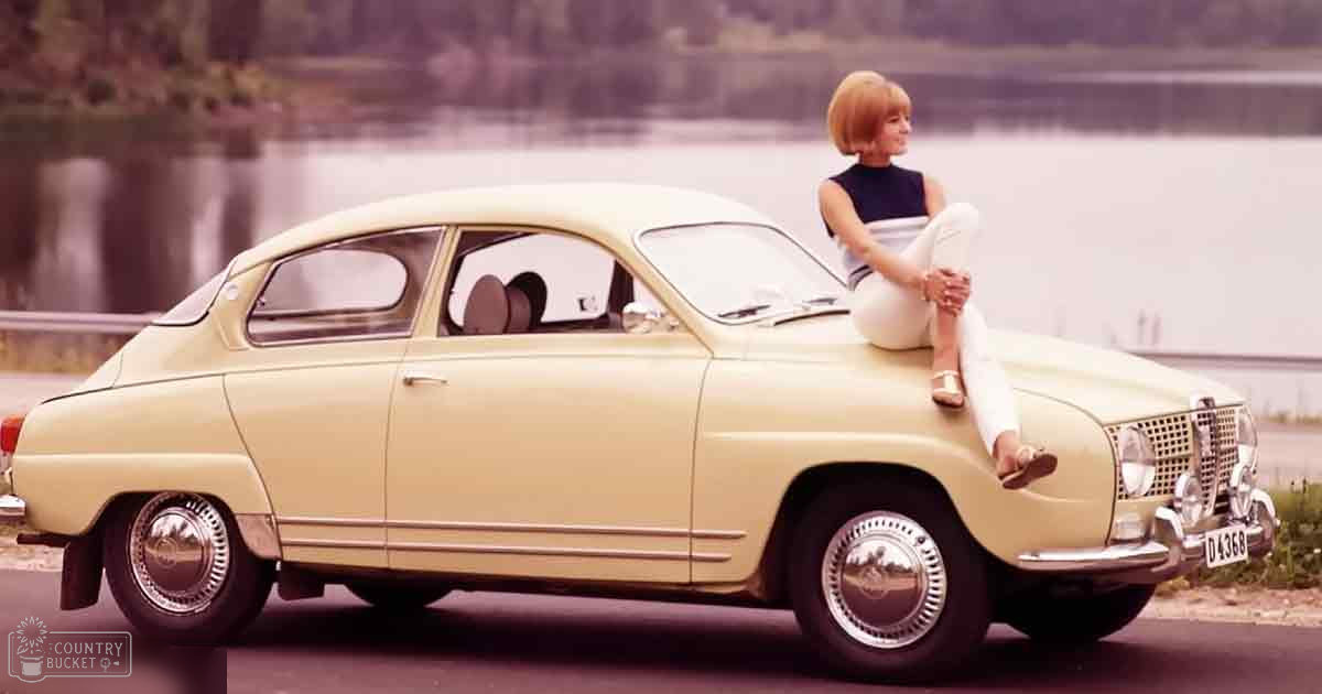 A young adult female sits on a vintage cream-colored Saab 96 car near a body of water, surrounded by trees and natural light.