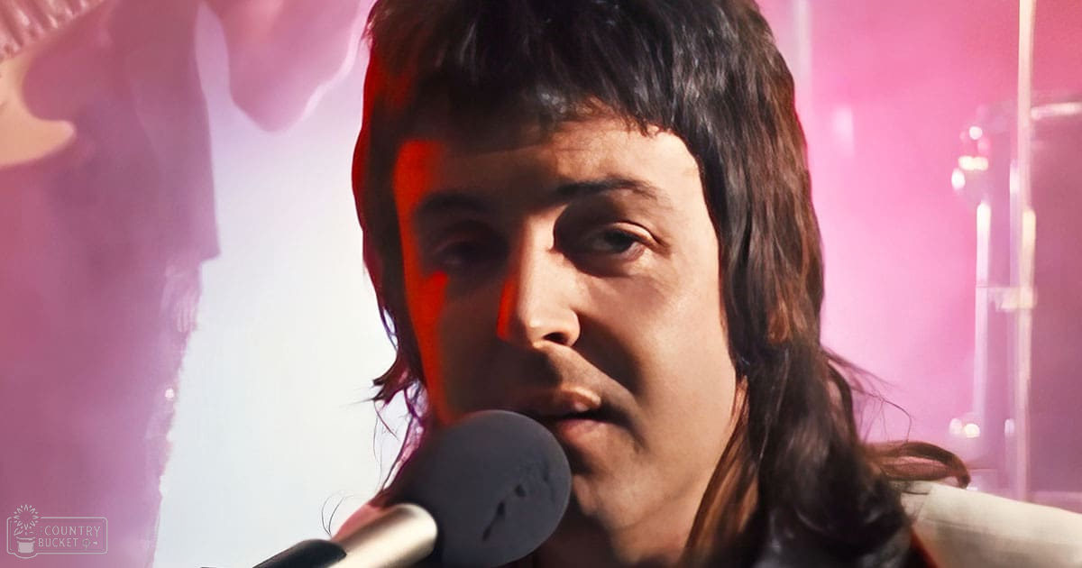 Adult male singer wearing a light-colored suit jacket performing on stage with pink lighting, microphone prominently displayed.