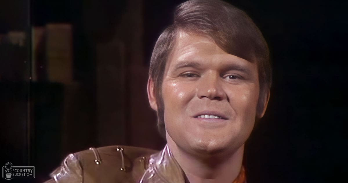 A middle-aged adult male wearing a shiny, metallic gold jacket with visible buttons or closures down the front and shoulder straps stands against dark walls in an indoor setting.