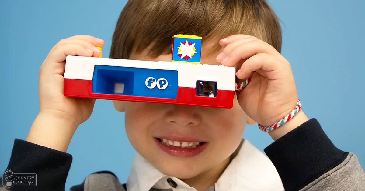 A toddler child wearing a white long-sleeved collared shirt with red, white, and blue accents holds a toy camera against a light blue backdrop.