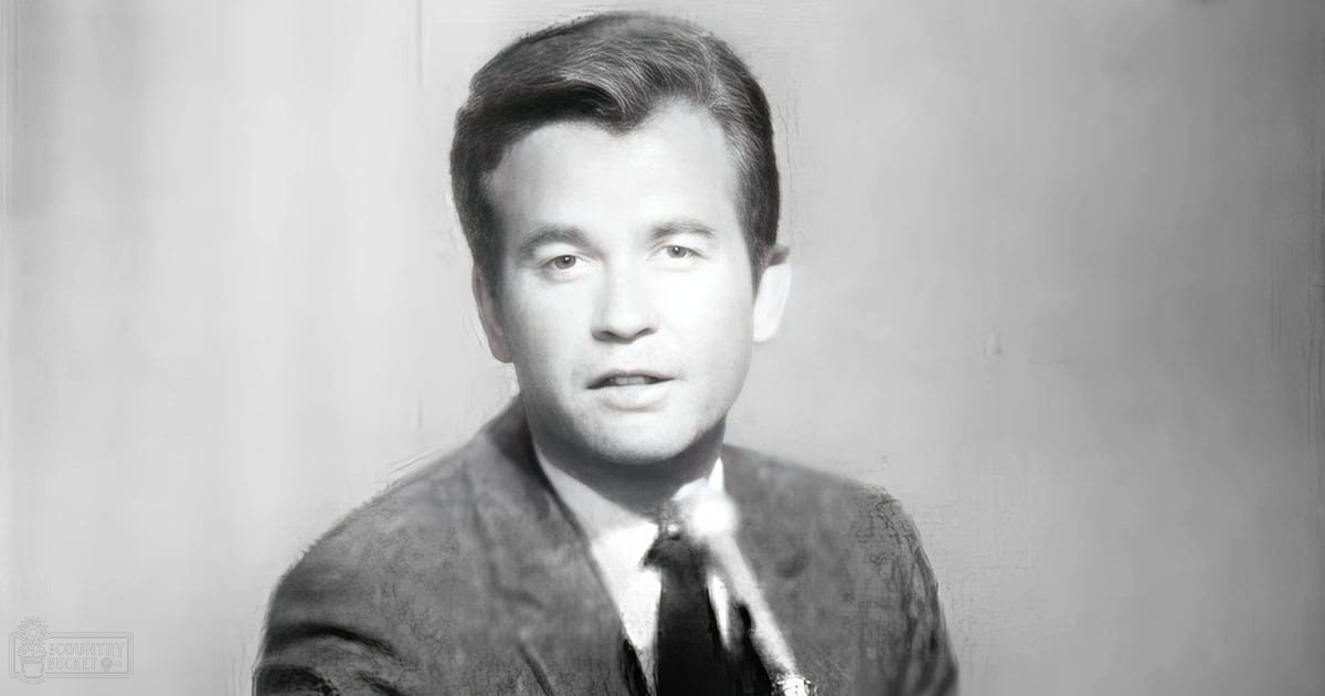 A young adult male in a formal suit jacket with a patterned texture, collared shirt, and long sleeves stands against an indoor background with plain wallpaper or paint.