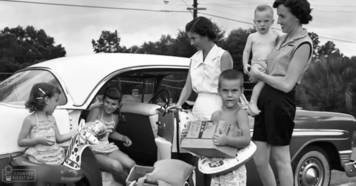 A family scene with children in colorful dresses and an adult holding a baby against the backdrop of a vintage car, trees, and power lines on what appears to be a summer day.