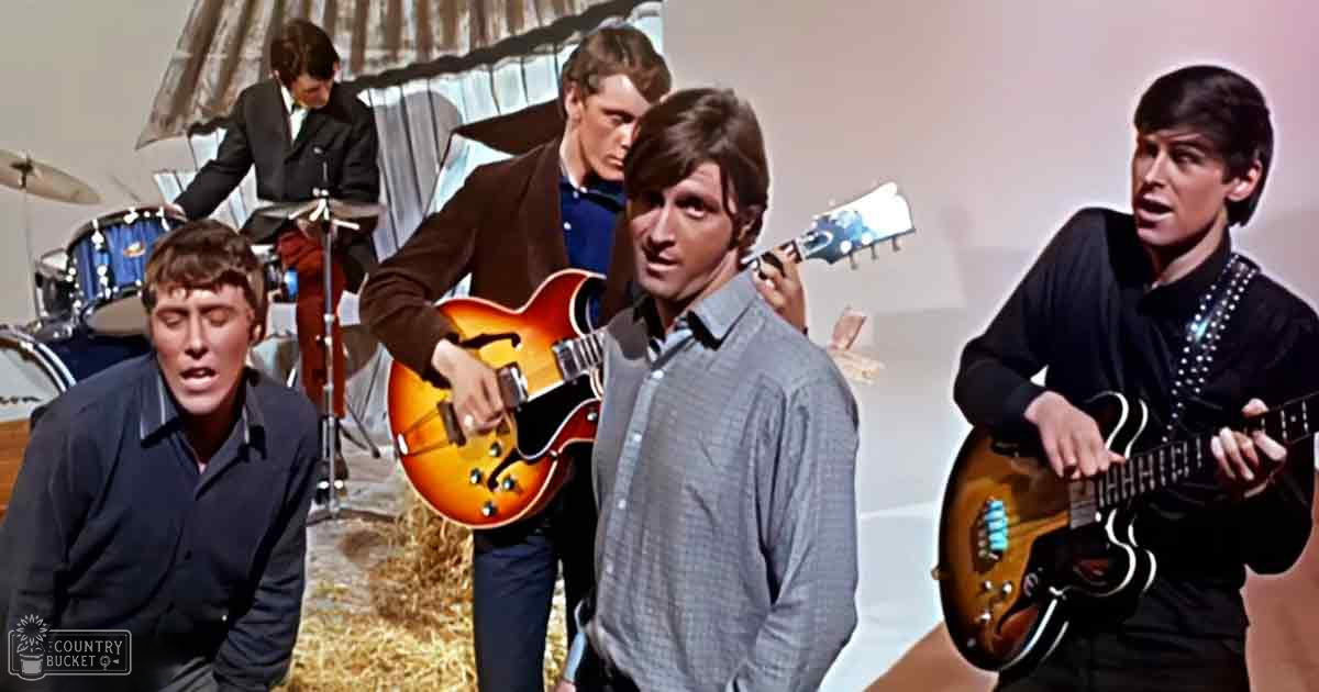 A musical performance scene from an older film or television show with young adult males in various attire, including a drummer in a dark suit jacket and a guitar player with a sunburst finish guitar. The setting appears to be an indoor stage or studio, possibly from the 1960s.