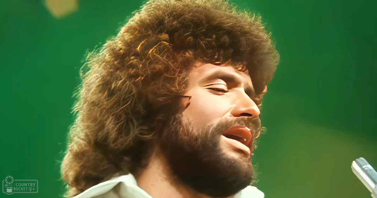 A performer singing in a concert setting with green background and stage lighting, wearing a white shirt from the 1970s or early 1980s.