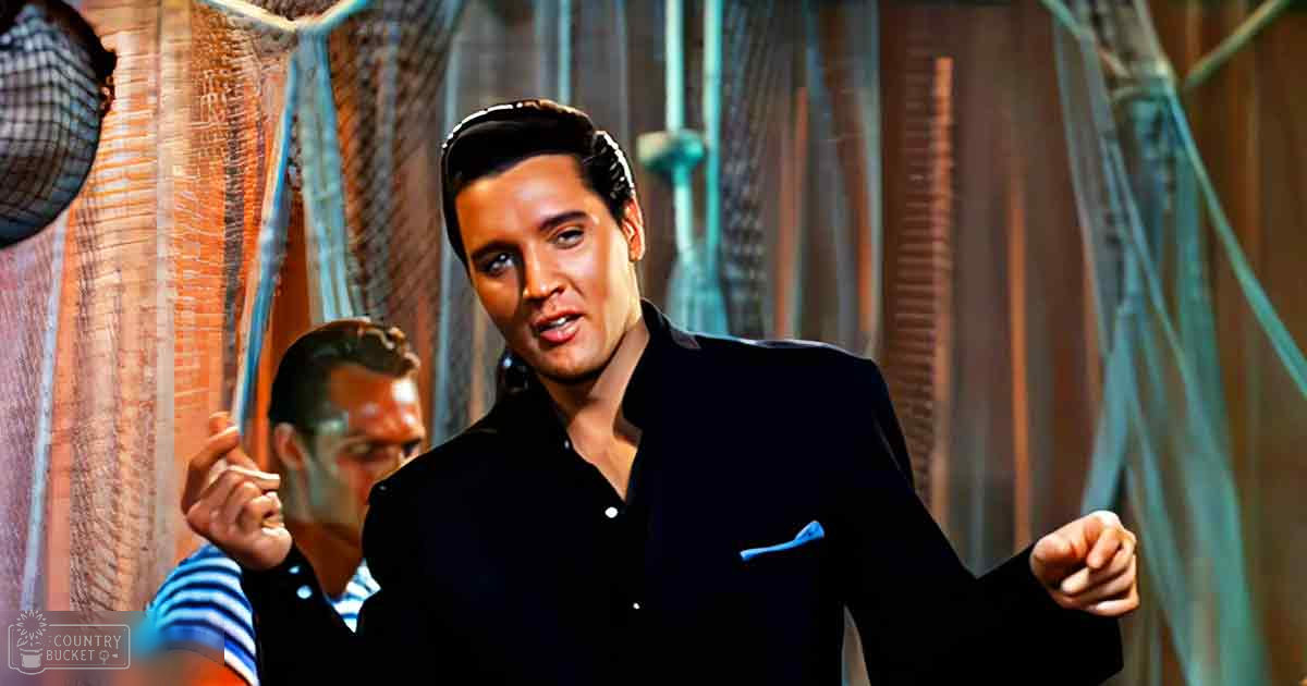 A man in his adult years, dressed in a black suit jacket with a blue pocket square and dark hair styled upwards, stands indoors against a backdrop of fishing nets or fabric. Another individual, wearing a striped shirt and vest, is visible nearby.