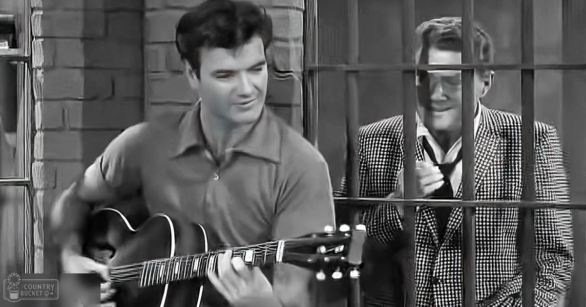 A young adult male playing guitar in front of another individual behind bars in what appears to be a jail setting.