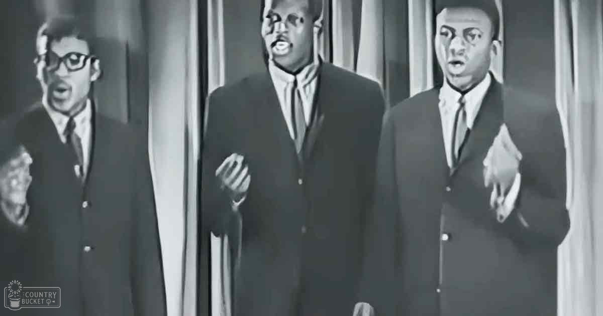 Adult males singing in front of a curtain indoors, likely at an event or performance setting.