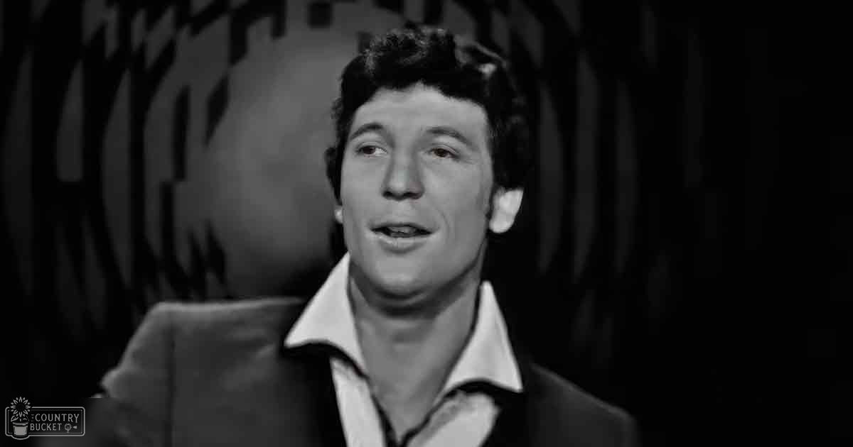 A young adult male in an indoor setting wearing a dark-colored suit jacket over a white collared shirt with the top buttons undone; lighting suggests it could be evening or night.