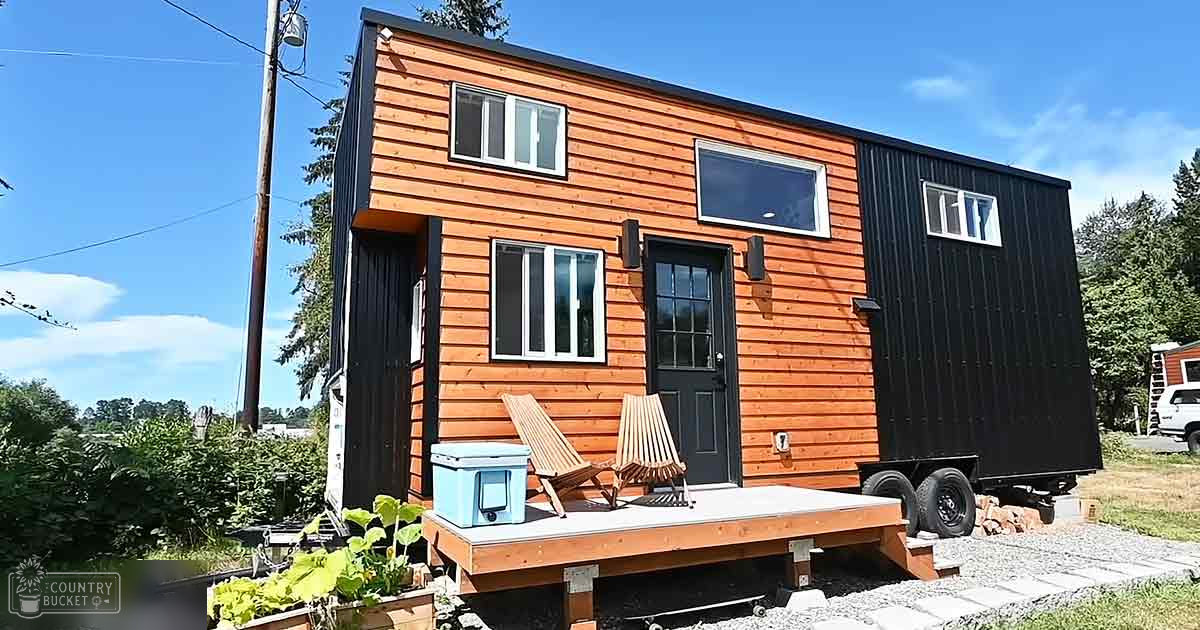 A tiny house with orange and black siding sits on a small deck, surrounded by greenery under a clear blue sky.