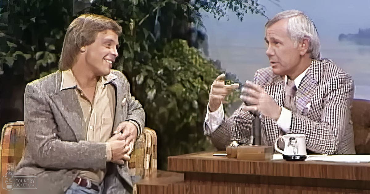 Talk show host interviewing an older male guest in a studio setting with patterned chairs and greenery visible through windows.