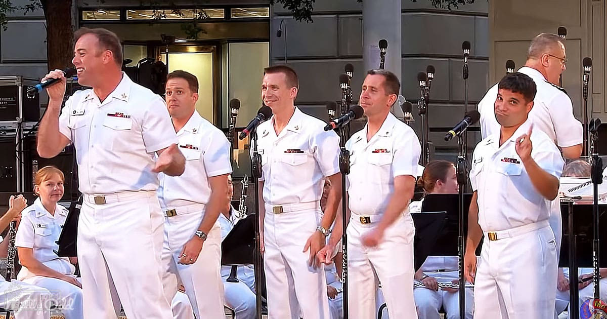 A group of men in white uniforms singing outdoors on a sunny day. Trees and buildings are visible in the background.