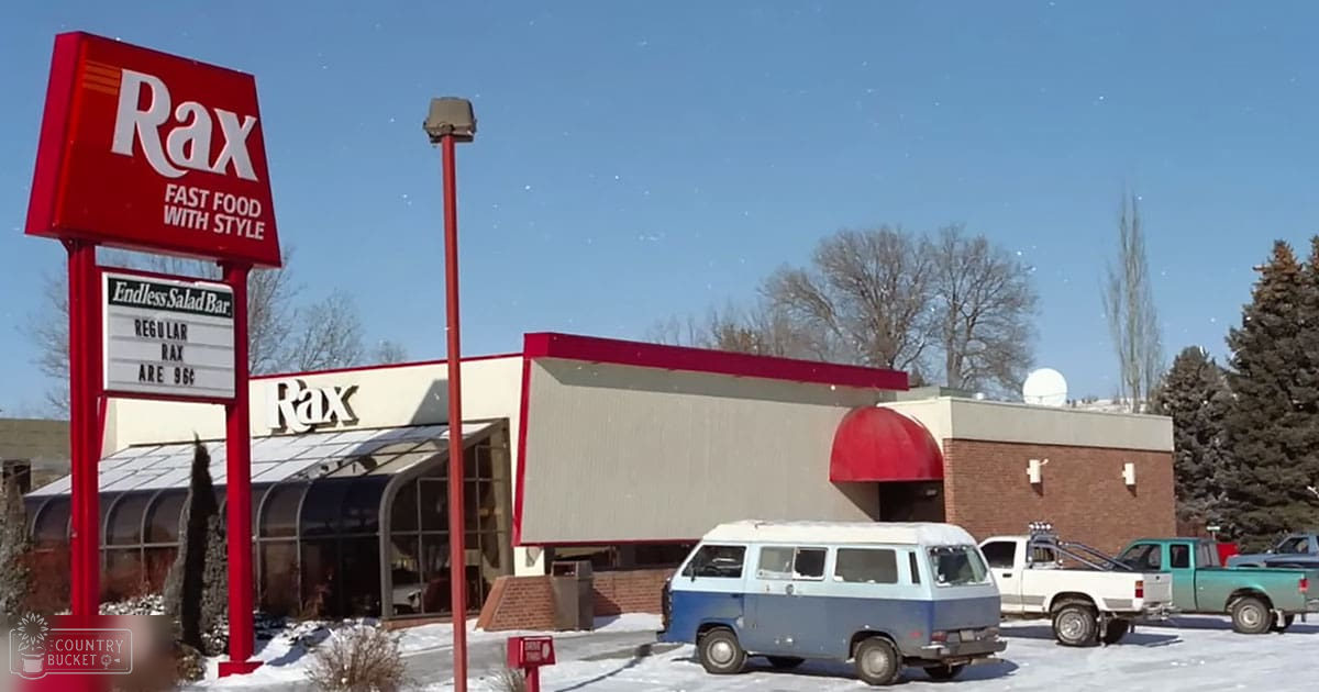 A clear winter day at a Rax fast food restaurant with snow on the ground, surrounded by trees without leaves and parked cars in front of the building.