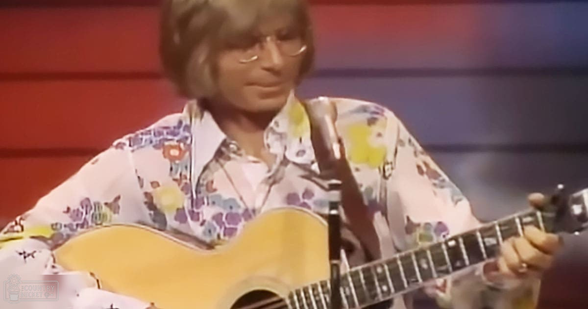 An adult male musician in a studio setting, wearing a loose-fitting long-sleeved floral-patterned shirt with vintage 1960s or early 1970s style, holding an acoustic guitar and wearing a necklace.