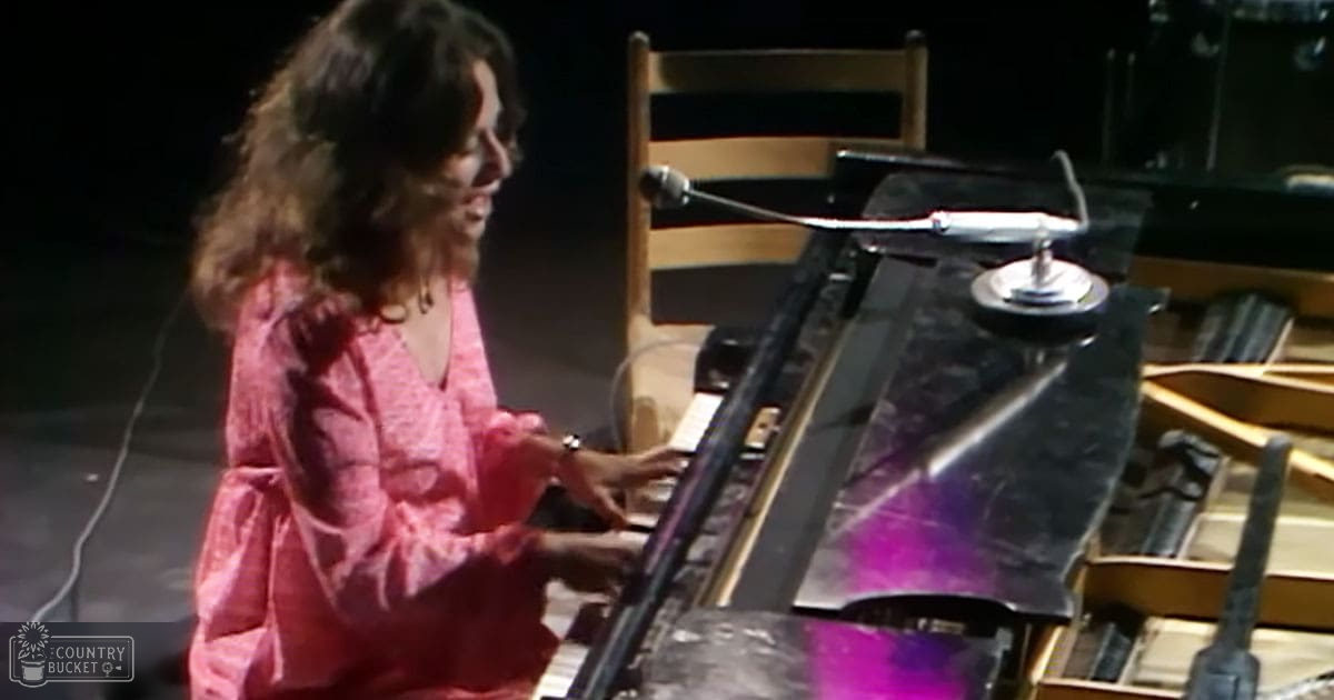 A woman in a pink dress plays a grand piano under dim evening light, surrounded by simple furniture.