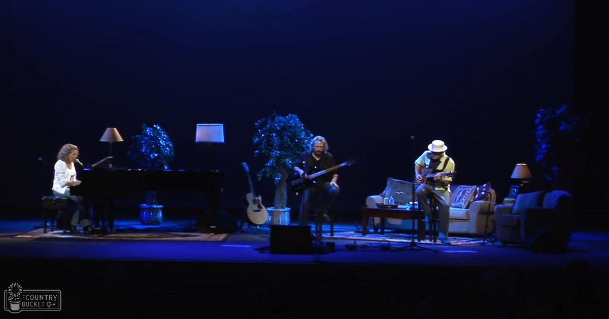A musical performance indoors with a grand piano, an acoustic guitar, and a bass guitar on stage under dim evening lighting.