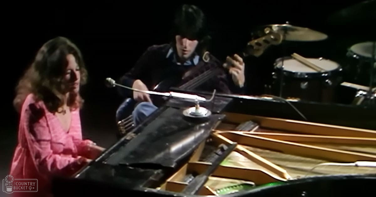 An indoor musical performance scene with two musicians on stage; a female singer in a pink jacket and male musician playing cello, both under controlled lighting.