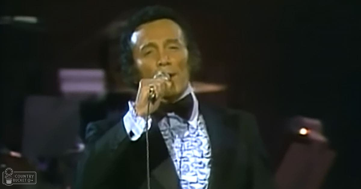 An adult male performer in a dark suit jacket over a patterned shirt with long sleeves slightly rolled up, holding a microphone on stage at a concert venue.