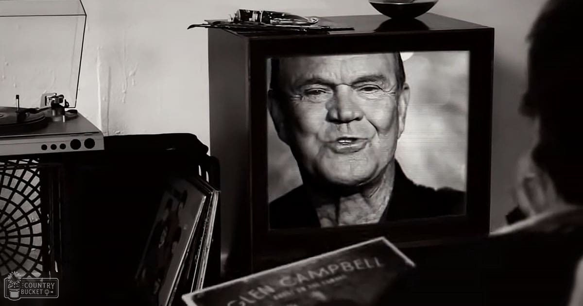 An older adult male in a dark-colored shirt or jacket, displayed on a television screen within an indoor home setting with minimal decor and muted colors.